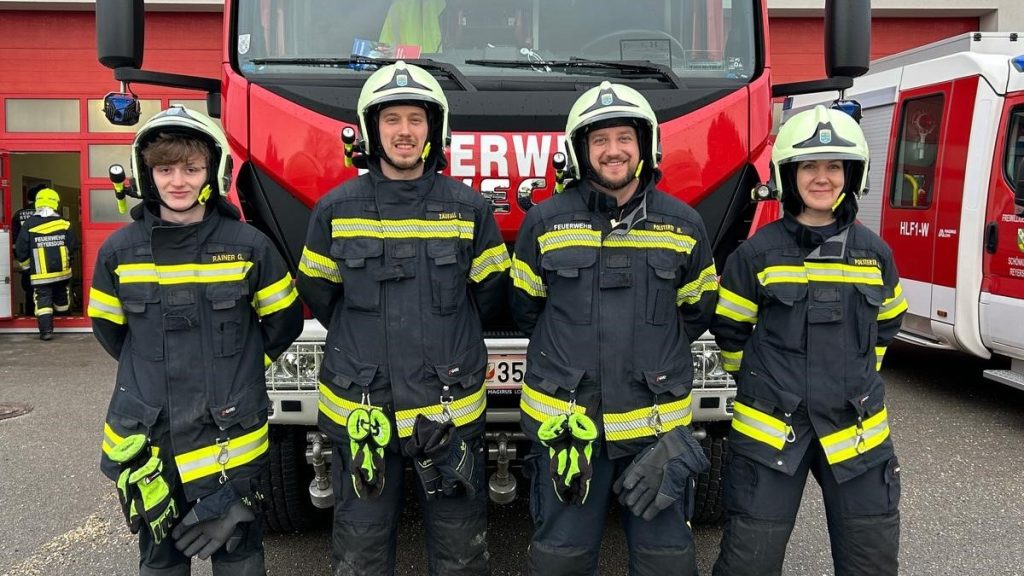 Vier Feuerwehrleute in kompletter Schutzausrüstung mit Helmen stehen lächelnd nebeneinander vor einem großen roten Einsatzfahrzeug, im Hintergrund weitere Feuerwehrfahrzeuge und Personen.