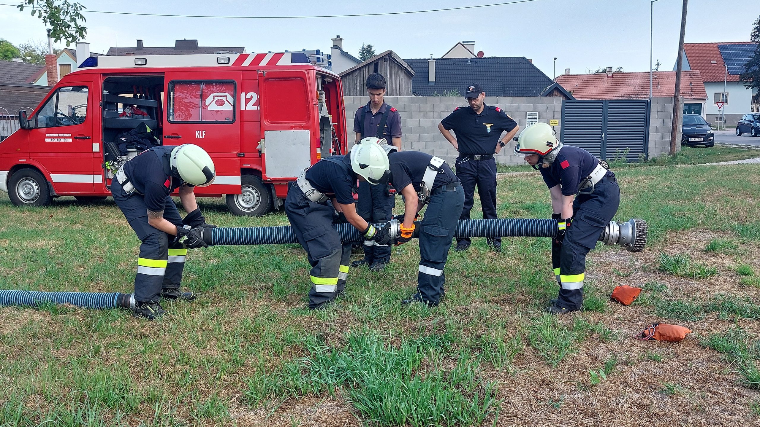 Vier Feuerwehrleute in Einsatzkleidung und Helmen kuppeln auf einer Wiese einen Saugschlauch zusammen, während zwei weitere in schwarzer Uniform das Geschehen beobachten; im Hintergrund steht ein geöffnetes rotes Feuerwehrfahrzeug.