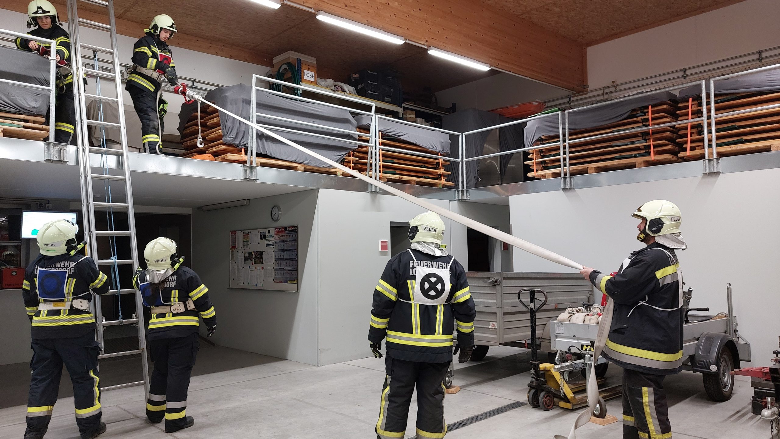 Mehrere Feuerwehrleute in Einsatzkleidung üben in einer Halle das Arbeiten mit Seilen, Leitern und einem Schlauch über zwei Ebenen; zwei Personen stehen auf einer Empore mit gestapelten Tischen, andere sichern unten mit Seil und Schlauch, eine Leiter steht bereit.