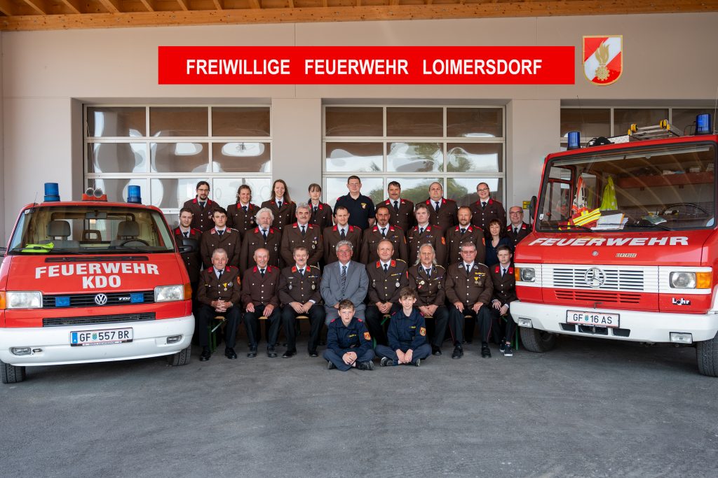 Gruppenfoto der Freiwilligen Feuerwehr Loimersdorf vor dem Feuerwehrhaus; etwa 28 Mitglieder in brauner Uniform, darunter auch Frauen und zwei Jugendliche in blauer Einsatzkleidung, eingerahmt von zwei roten Feuerwehrfahrzeugen mit der Aufschrift „FEUERWEHR“.
