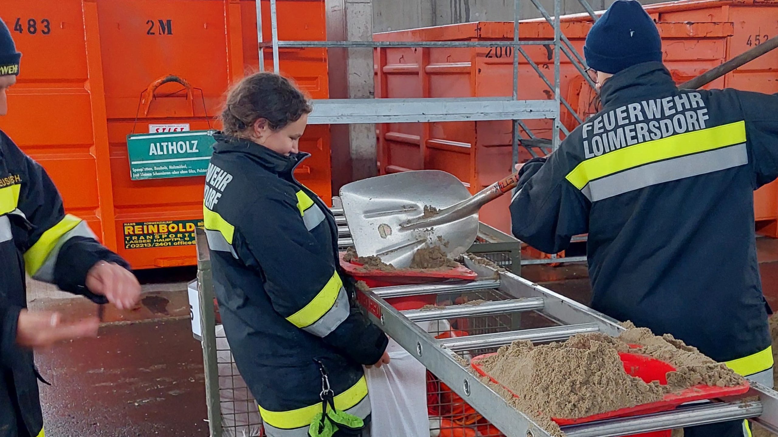 Mitglieder der Feuerwehr Loimersdorf in wetterfester Einsatzkleidung befüllen bei einer Sandsackaktion Schaufel für Schaufel Sandsäcke; im Hintergrund orange Container mit der Aufschrift „Altholz“.